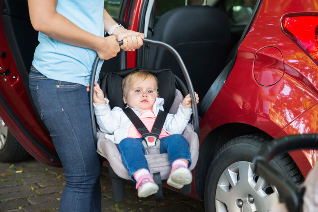 Une personne tient une coque bébé devant une voiture dont la portière est ouverte